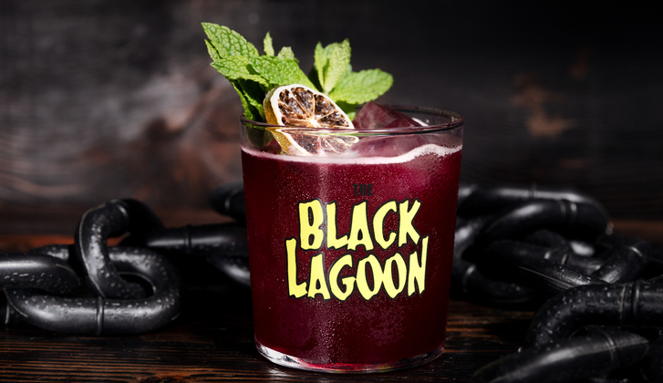 Close up of dark maroon cocktail with mint and blood orange garnish that says Black Lagoon on glass