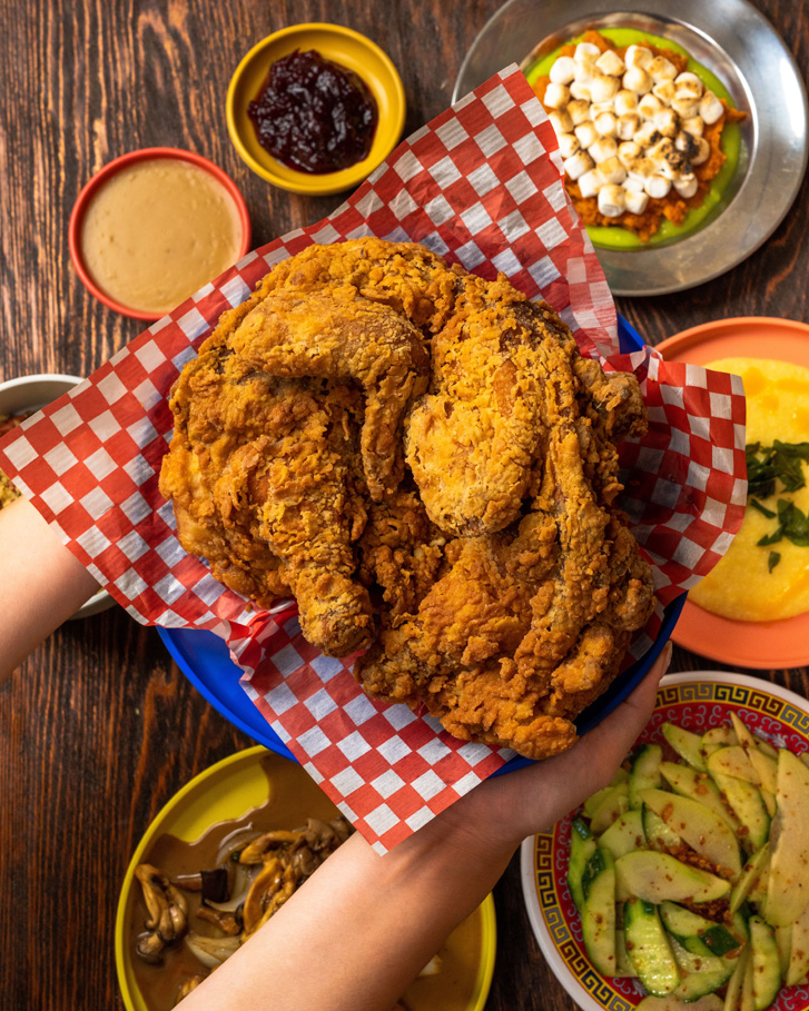 Hands holding fried chicken over top a table of potatoes and sides