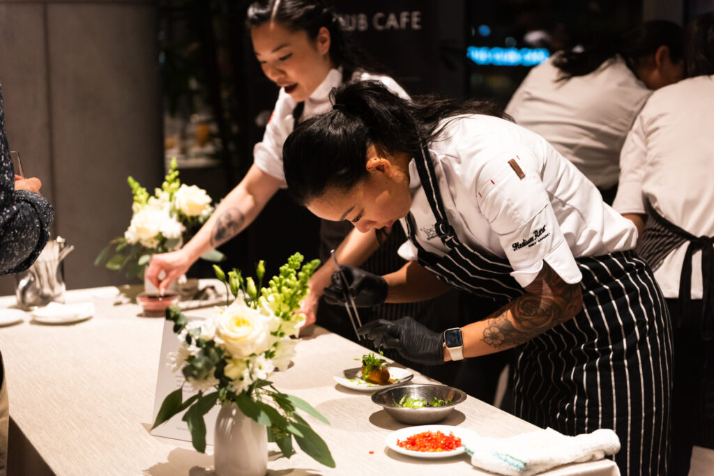 Two chefs in white shirts and aprons prepare tasting plates.