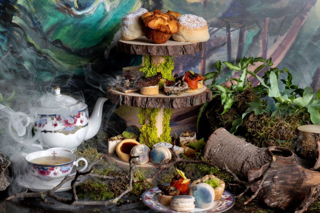 Table with tea pot and afternoon tea food display, with wood and forest decorations in front of a painted background