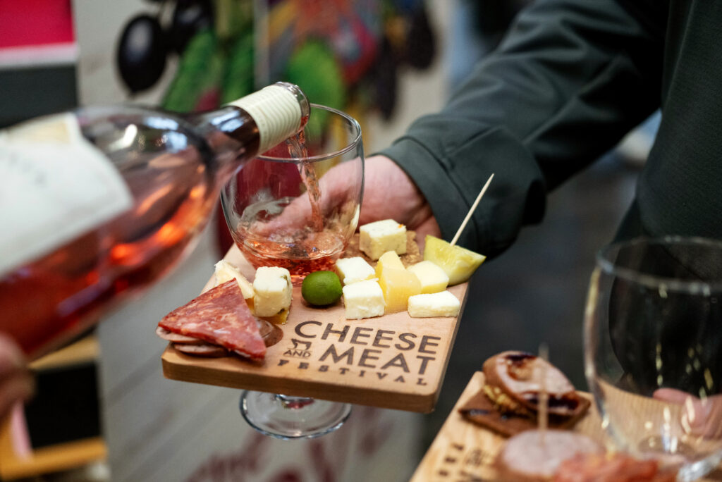 Someone pours wine into a glass alongside someone holding a plate of meat and cheese. 