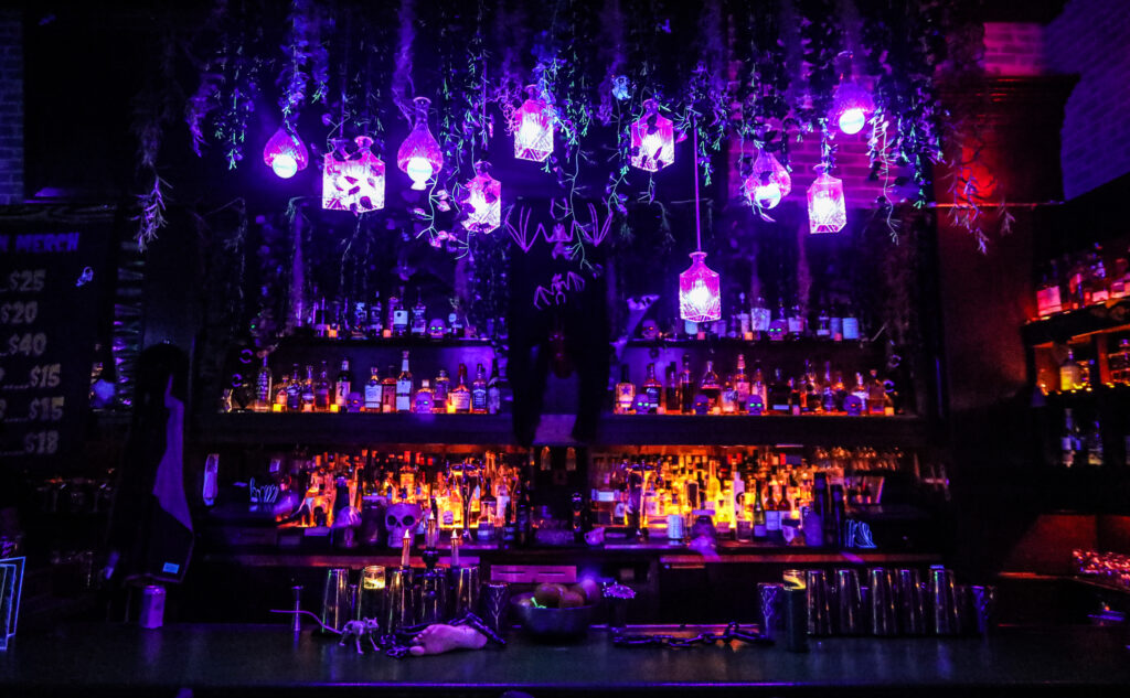 A spooky dark bar set up with purple lights and lightly glowing alcohol bottles lining the walls. 