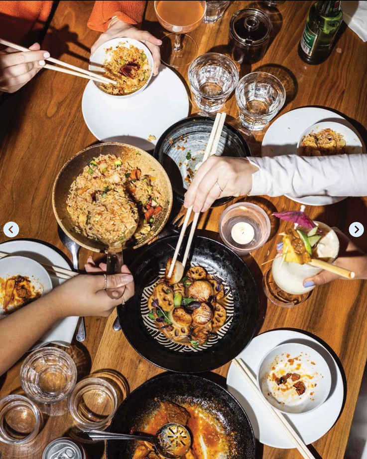 hands reaching across a table with chopsticks for food from a variety of dishes