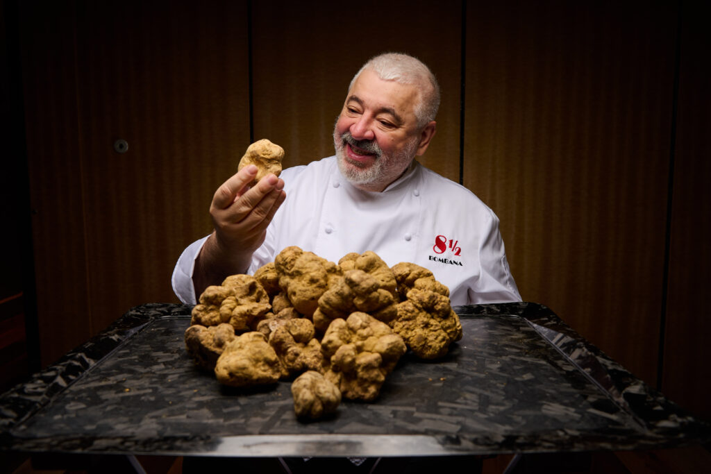 A chef holds up a large white truffle mushroom over a tray of mushrooms.