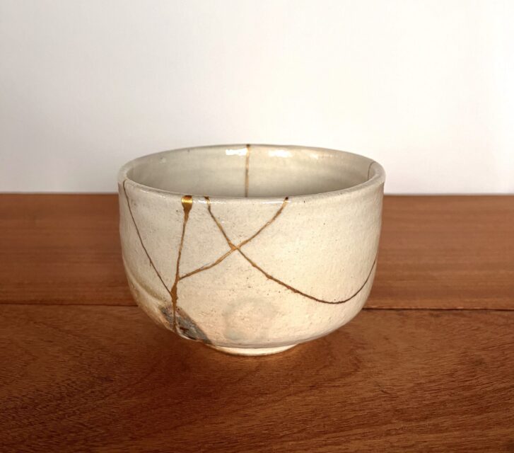 A ceramic bowl repaired with the Japanese art of Kintsugi, featuring gold-filled cracks that highlight its once broken parts. The bowl rests on a wooden surface with a plain background, showcasing the beauty in its imperfections.