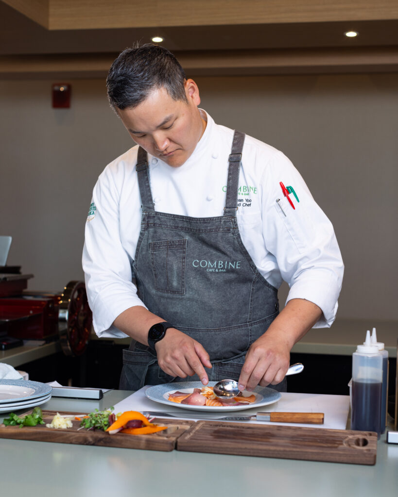 A chef in a white shirt and blue apron plates a dish artfully. 