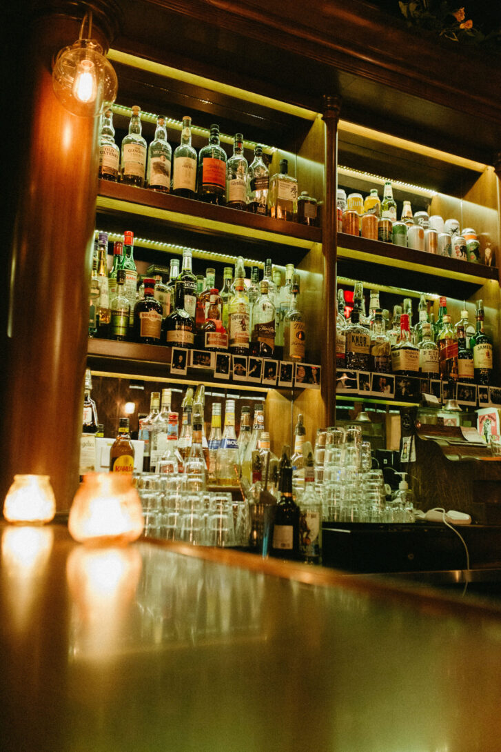 Close-up of a well-stocked bar with shelves filled with a variety of liquor bottles, including whiskey, gin and rum. The warm lighting highlights the bottles, creating a cozy and inviting atmosphere. Glassware is neatly arranged below the shelves, ready for use.