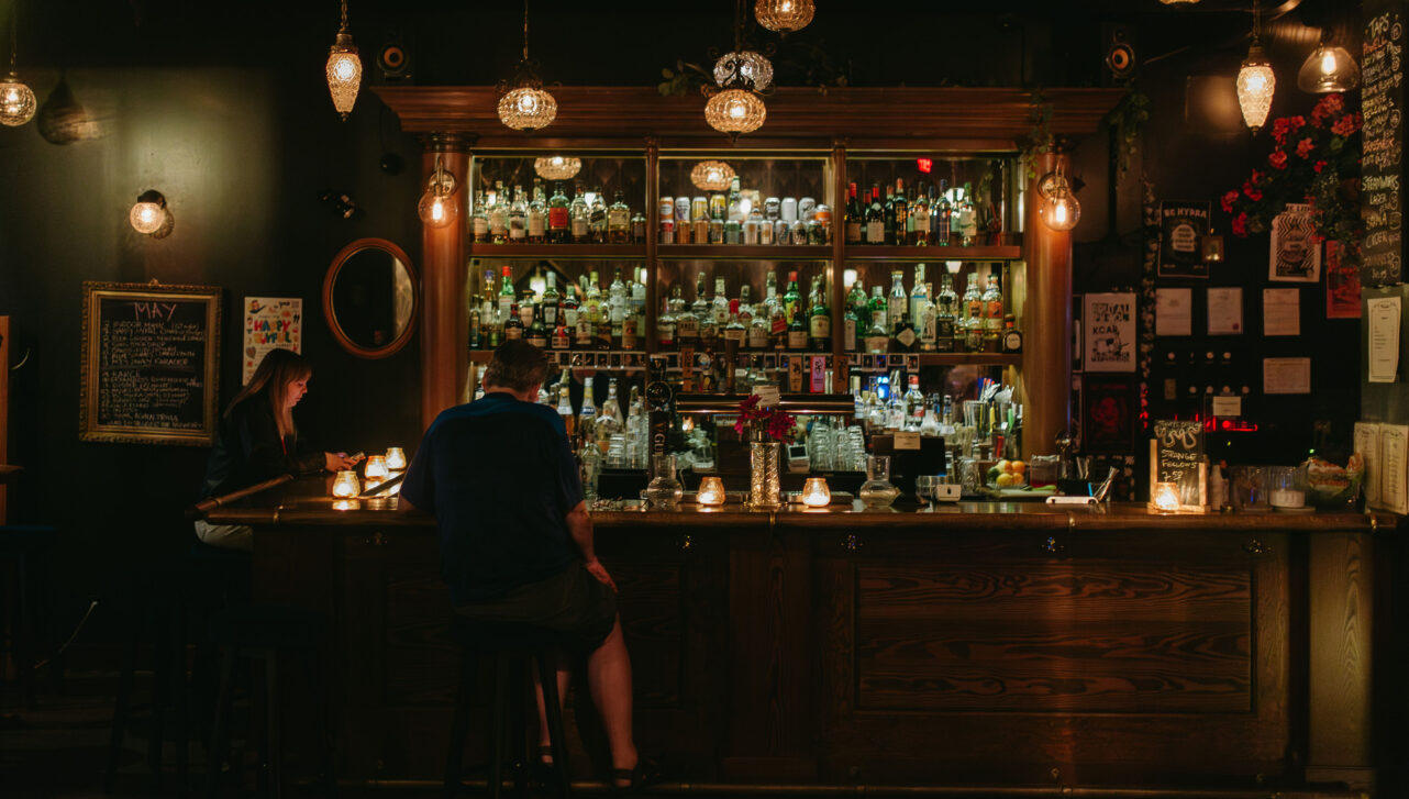 A dimly lit bar with warm lighting, featuring a wide selection of bottles on shelves behind the bar. Two patrons are seated at the bar, engaged in quiet conversation. The bar is adorned with vintage light fixtures and a small chalkboard menu on the wall.