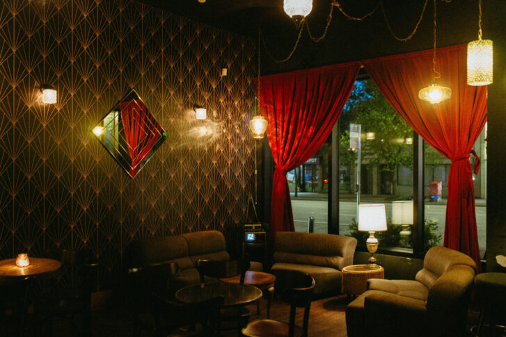 Cozy lounge area with a vintage ambiance, featuring dark patterned wallpaper, red velvet curtains and warm lighting from hanging lamps and wall sconces. Comfortable sofas and chairs are arranged around small tables, creating an inviting space for relaxation and conversation.
