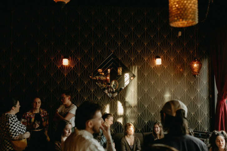 A group of people socializing in a dimly lit lounge with elegant, patterned wallpaper. Warm lighting fixtures and decorative wall mirrors create a cozy and inviting atmosphere, perfect for an intimate gathering or night out.
