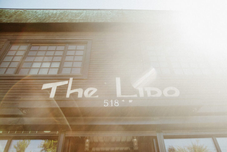 A sunny view of the entrance to The Lido, featuring its distinctive sign and address '518.' The wooden facade and large windows hint at the cozy, inviting atmosphere within, while sunlight creates a warm, welcoming glow.