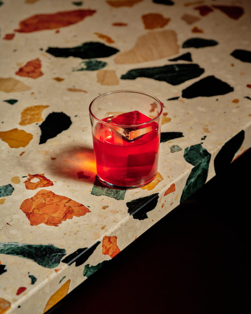 A negroni cocktail in a glass with an ice cube sits on a countertop. 