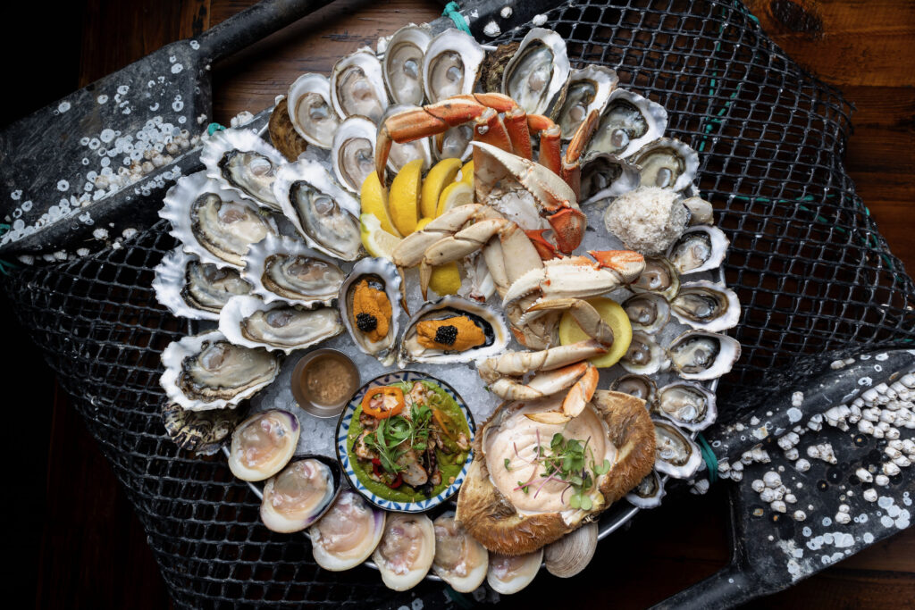 A large round platter of raw seafood including mussels, clams, oysters, prawns, and fresh lemon. 