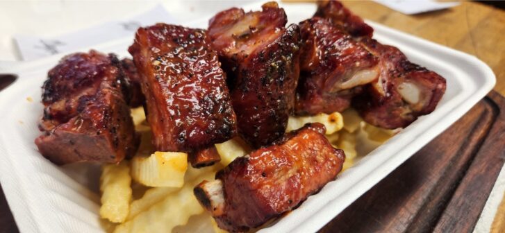 A close-up shot of glazed rib bites served on a bed of crinkle-cut fries in a white disposable container.