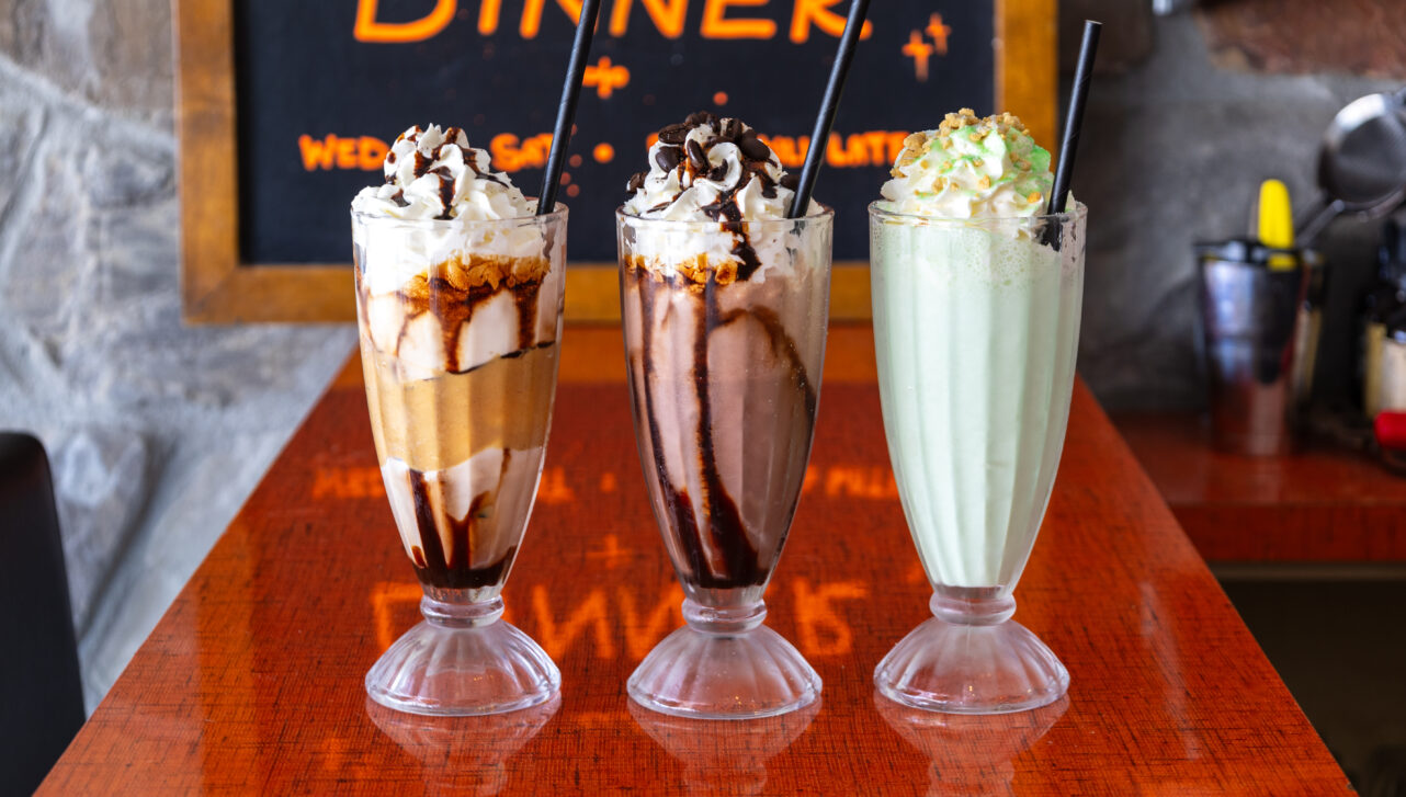 Three milkshakes topped with whipped cream on a red countertop.