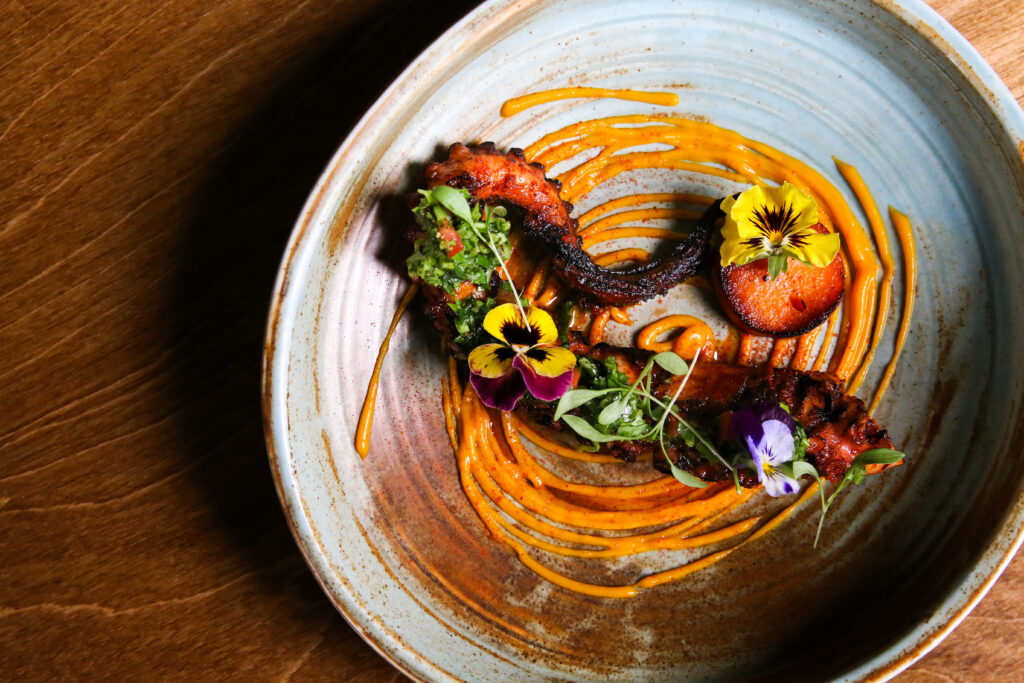 A dish featuring octopus garnished with fresh flowers and a swirl of orange sauce.