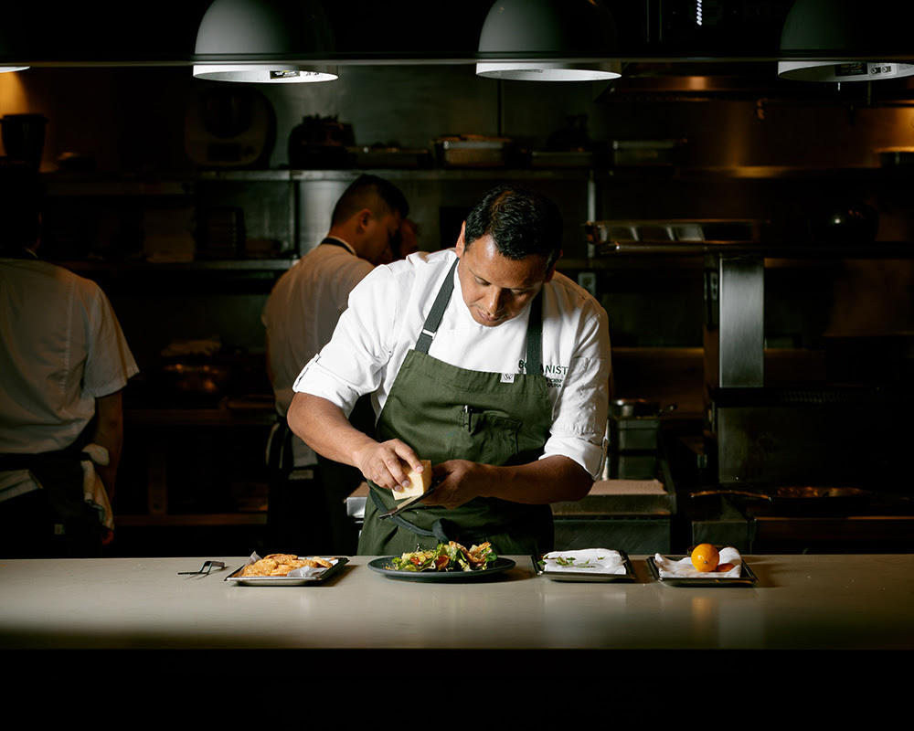 A chef in a white shirt and dark apron plates a dish against a moody dark backdrop.