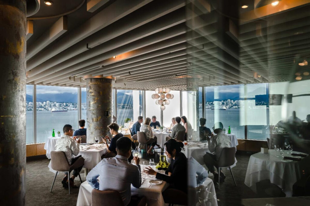 Finely dressed people dine in a large dining room with views of the ocean and mountains beyond.